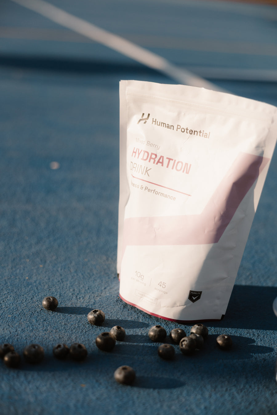 A stand-up pouch of Human Potential Hydration Drink Mix stands on a blue running track surface with scattered blueberries in the foreground.