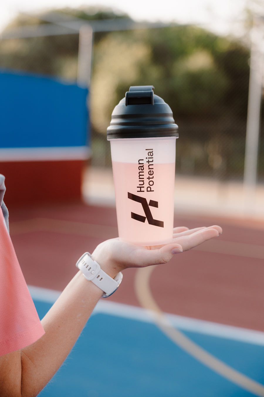 A person holds a translucent pink shaker bottle with a black lid on an outdoor sports court. The bottle features the text 'Human Potential' and a stylized logo.
