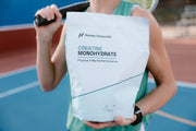 A person on a tennis court holds a large white bag of Human Potential Creatine Monohydrate. The person is wearing a green athletic tank top and holding a tennis racket, with the blue and red tennis court visible in the background.