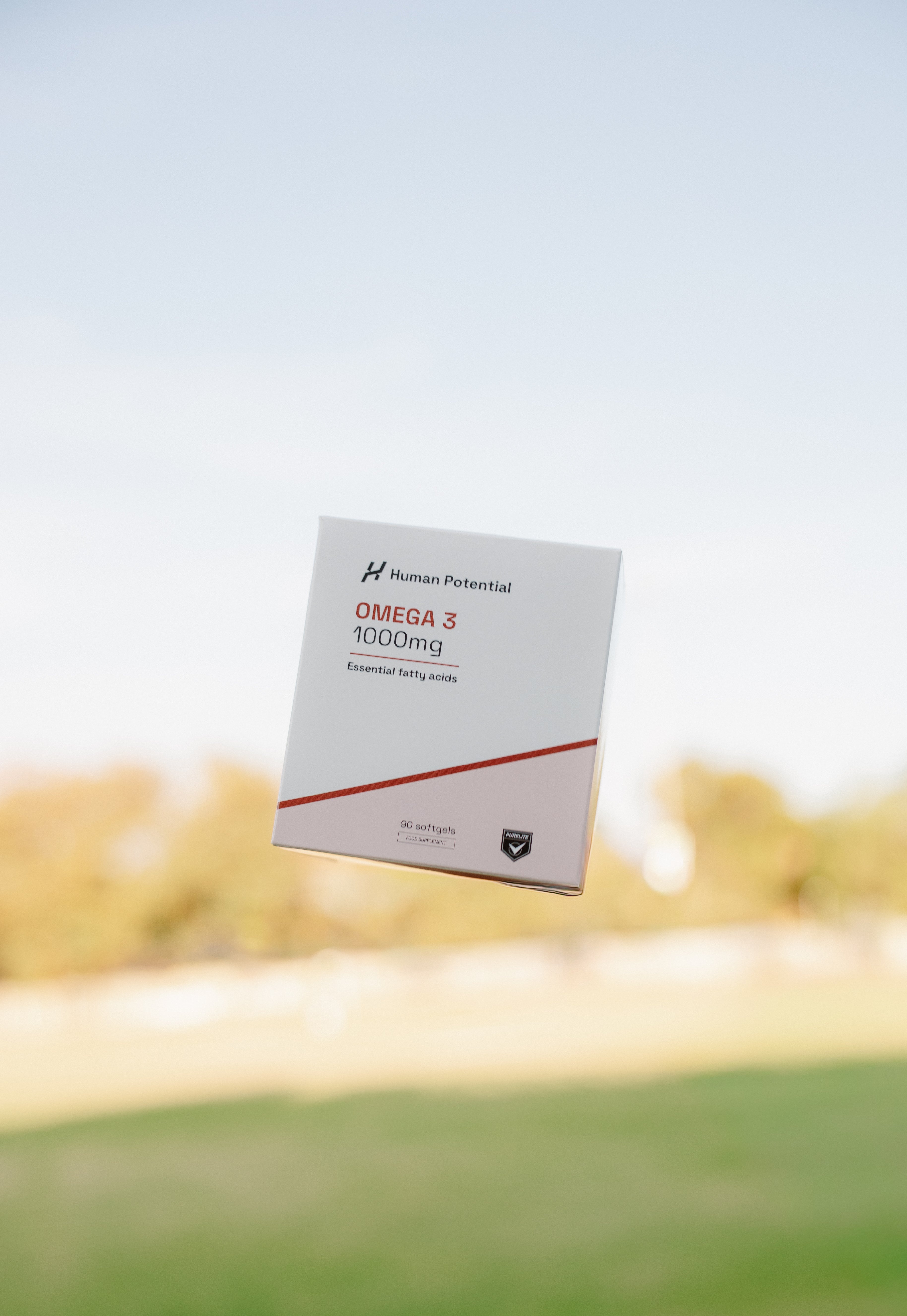 A boxed supplement product labeled 'Human Potential OMEGA 3 1000mg' floating against a blurred outdoor background of grass and trees under a pale blue sky.