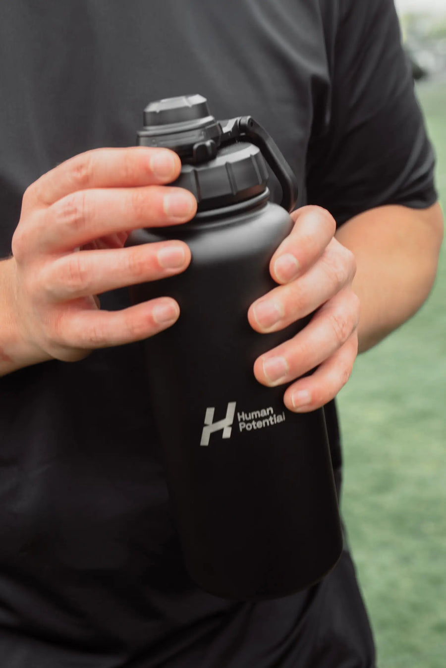 Person holding a black water bottle with 'Human Potential' logo against a blurred outdoor background