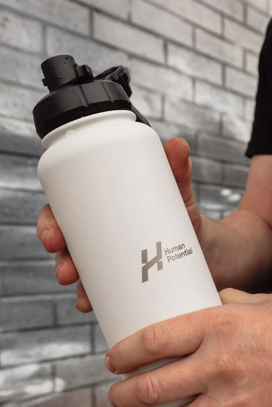 White water bottle with black cap held by a person against a brick wall.