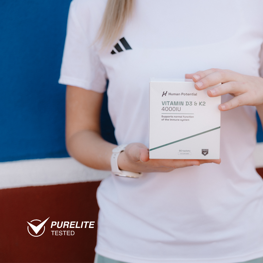 A person in a white Adidas T-shirt holds a white box of Human Potential Vitamin D3 & K2 4000IU. The background is a blue and brown wall, and a 'PURELITE TESTED' badge is in the bottom-left corner.